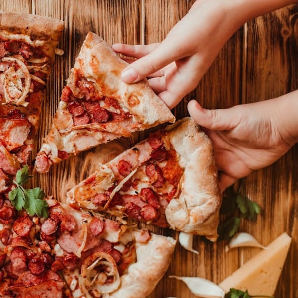 Hands taking slices of Bacio Pizza with sausage, onions, and cheese on a wooden table.