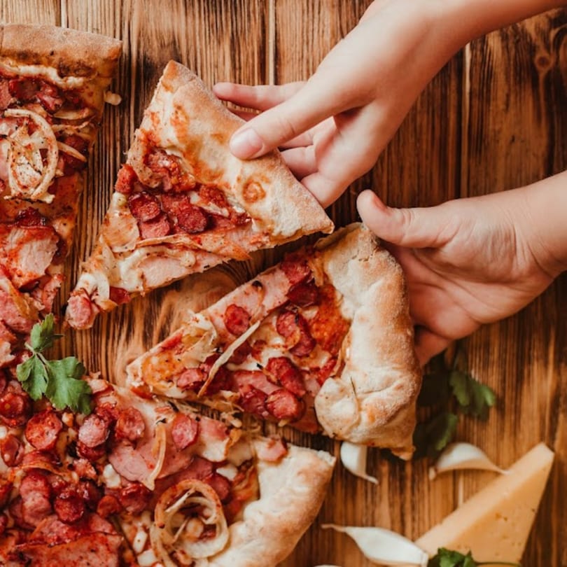 Hands taking slices of Bacio Pizza with sausage, onions, and cheese on a wooden table.