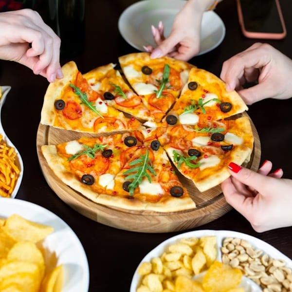 Hands reaching for slices of Bacio Pizza with olives and arugula, surrounded by snacks.