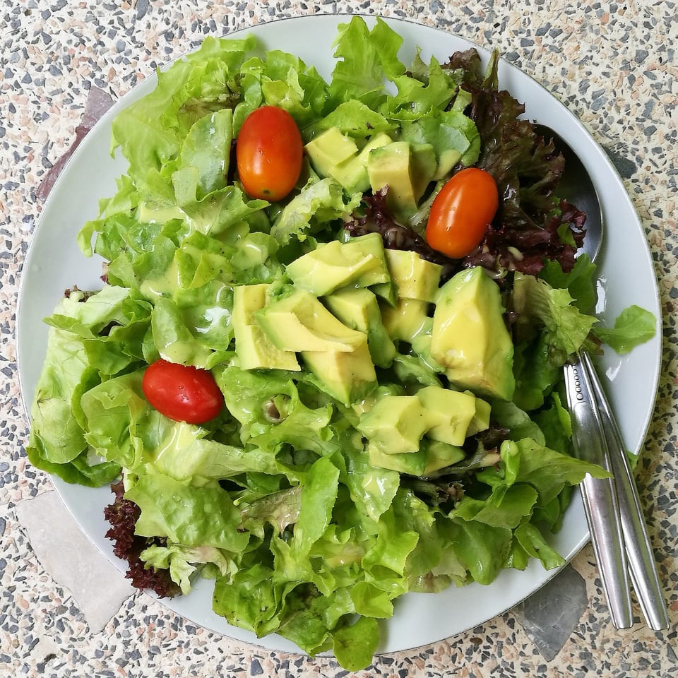 Fresh green salad with avocado chunks and cherry tomatoes on a white plate, with a fork and spoon.